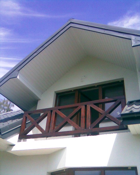 Balkon z drewnianą balustradą o wzorze krzyżowym, widok z dołu na tle błękitnego nieba.